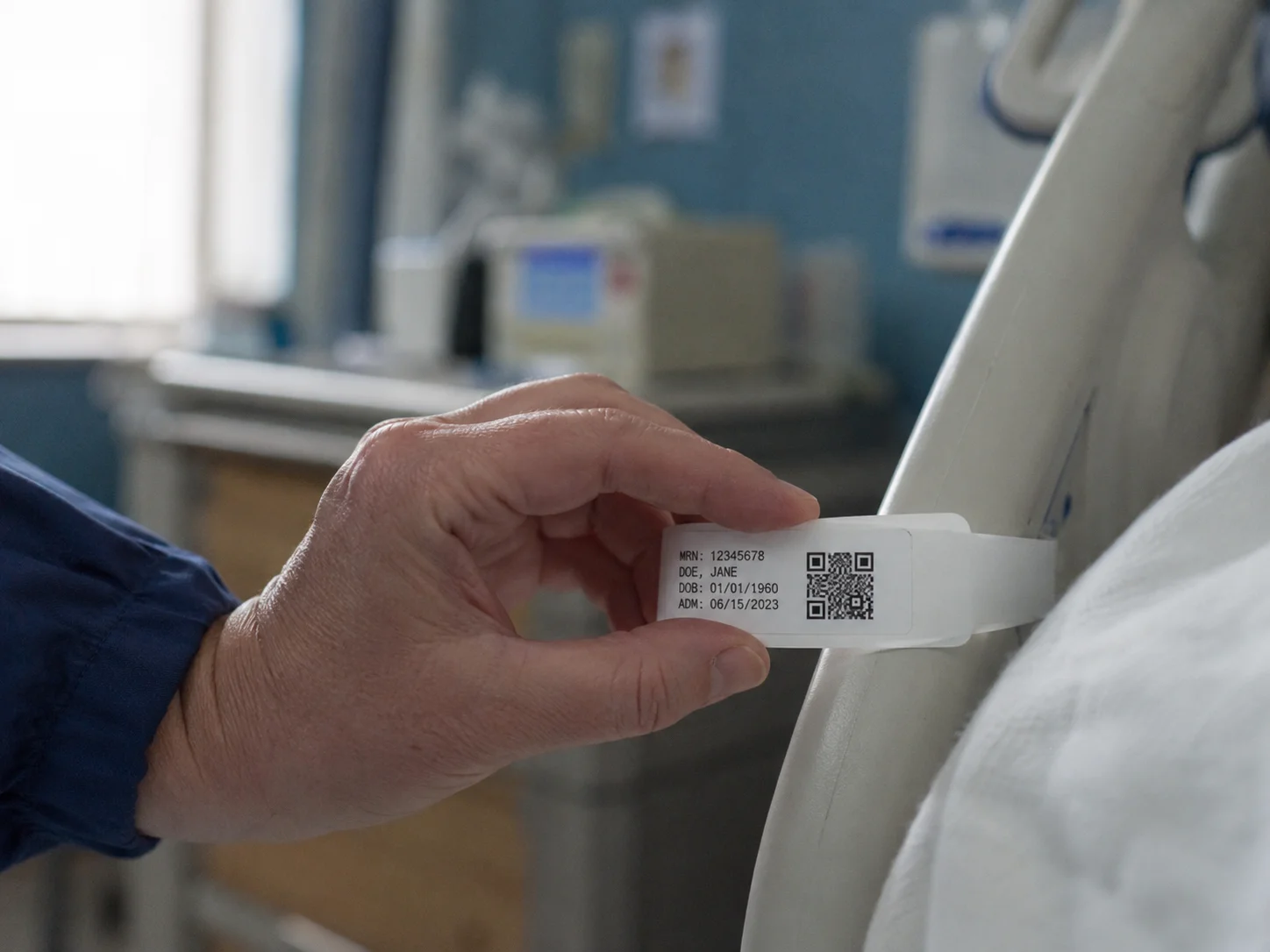 Hospital nurse holding a patient wristband with a printed QR code at a hospital bed rail in a clinical setting